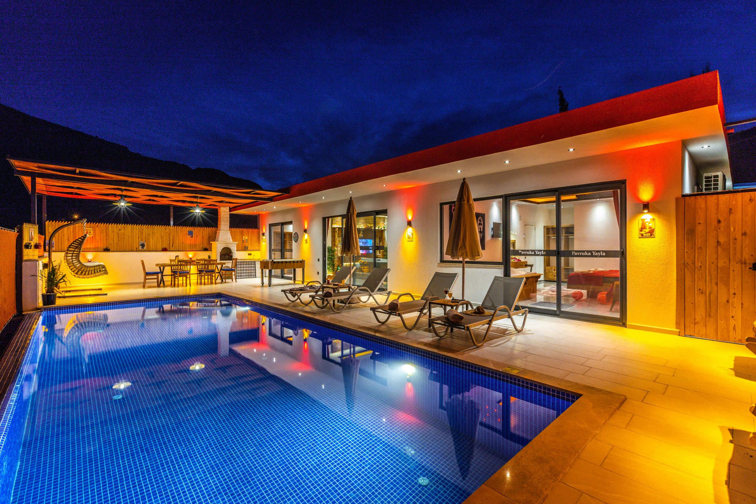 Modern villa with illuminated poolside area and deckchairs under night sky.